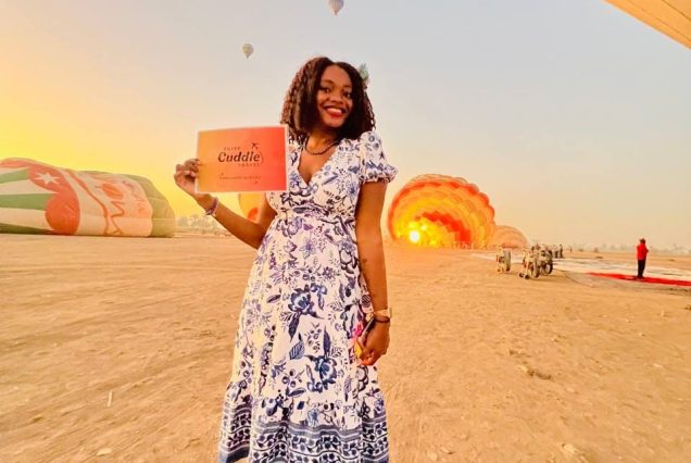 luxor hot air ballon : A vertical shot of a smiling female tourist standing at a hot air balloon launch site in Luxor at sunrise, holding a bright yellow "Cuddle Travel" sign in the foreground. In the background, numerous colorful hot air balloons are ascending against a glowing golden dawn sky over the desert landscape. The tourist is wearing a dark green dress and looking back over her shoulder with a joyful expression.