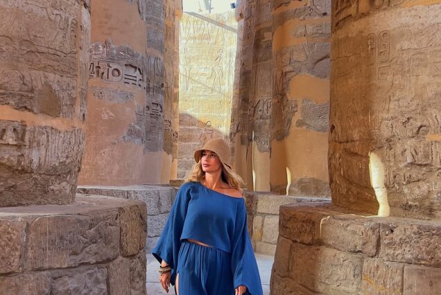 A wide-angle view of the Great Hypostyle Hall at Karnak Temple, featuring rows of massive sandstone columns carved with intricate hieroglyphics under a bright blue sky. Sunlight filters through the stone ruins, highlighting the weathered textures of the ancient pylons