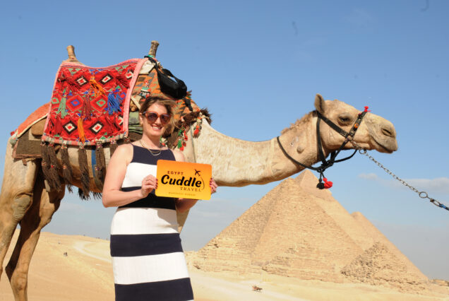 A happy traveler holding a "Cuddle Travel" brand sign at the Giza Plateau, with the Great Pyramids and a decorated camel in the background under a sunny sky.
