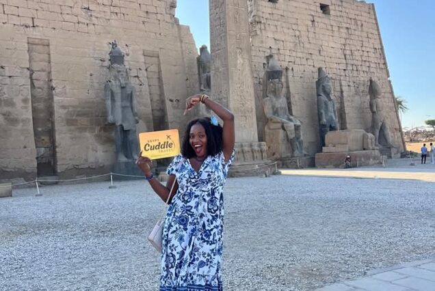 A woman in a blue and white dress standing in front of the massive pylon of Luxor Temple, featuring ancient statues of Ramses II and a towering obelisk. Karnak and Luxor Temple Private Tour