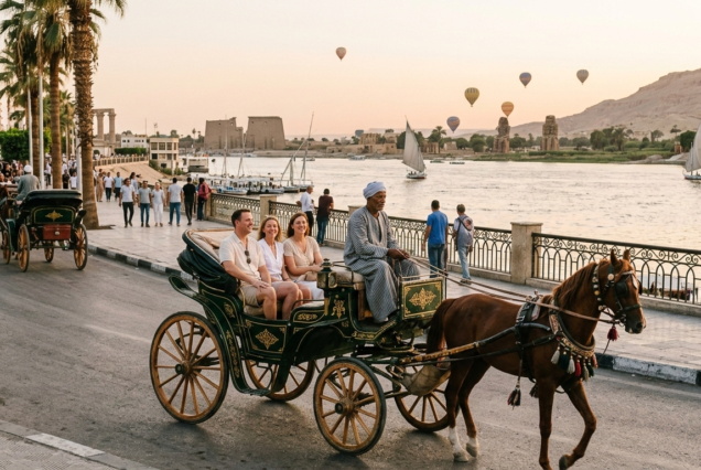 Karnak & Luxor Temple Private Tour: A digital illustration of a traditional green horse-drawn carriage (Hantour) carrying tourists along the Nile Corniche in Luxor at sunset, with hot air balloons and ancient monuments in the background.