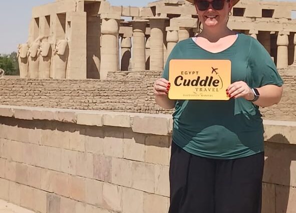 A woman standing in front of the Luxor Temple pylon featuring colossal statues of Ramses II and a towering obelisk.