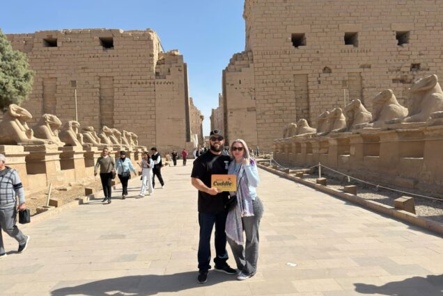 A couple holding a "Cuddle Travel" sign standing on the paved path of the Avenue of Sphinxes, flanked by ram-headed sphinx statues leading to the massive pylons of Karnak Temple.