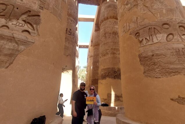 A tourist stands among the massive, towering sandstone columns of the Great Hypostyle Hall at Karnak Temple, covered in deep hieroglyphic carvings.