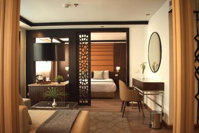 A spacious luxury cruise suite featuring a separate living area with a sofa, a wooden partition with an intricate geometric cutout, and a bedroom visible in the background.