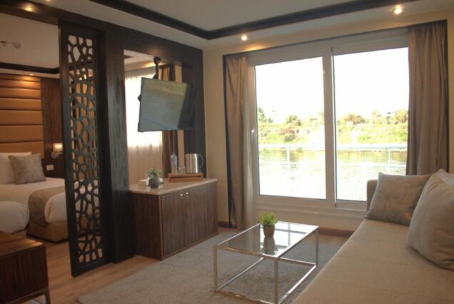 A modern suite living room with a large glass window showing the Nile riverbanks, a grey sofa, glass coffee table, and a decorative wooden screen.