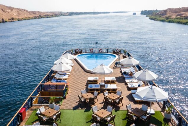 5 Days Nile Cruise from Luxor to Aswan |Abu Simbel A high-angle view of a cruise ship sun deck sailing on the Nile, featuring a blue triangular pool, lounge chairs with white umbrellas, and wooden dining tables.