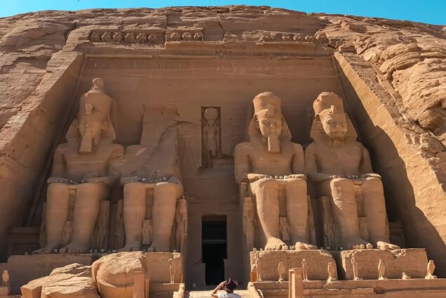 The grand exterior of the Great Temple of Abu Simbel, featuring four massive seated statues of Pharaoh Ramesses II carved into the sandstone cliffside under a bright sun.