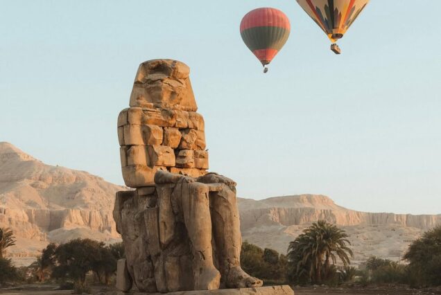 A giant ancient stone statue of Amenhotep III (Colossi of Memnon) in Luxor, with two colorful hot air balloons floating in the clear morning sky and the Theban mountains in the background Luxor West Bank Private Tour